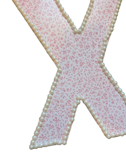 A decorative letter X cutout with a pink and white floral pattern, bordered with white faux pearls, displayed on a gray wood floor.